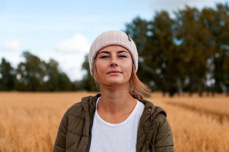 年轻的深色头发的女人，戴着一顶针织的粉红色帽子，来自一种天然毛皮，在秋天的一天，在一片金色的田野和一片森林的背景下微笑摆着姿势。