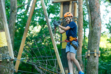 快乐可爱的小男孩穿着蓝色T恤和橙色头盔，在冒险公园玩，拿着绳子和爬木楼梯。 积极的生活方式概念