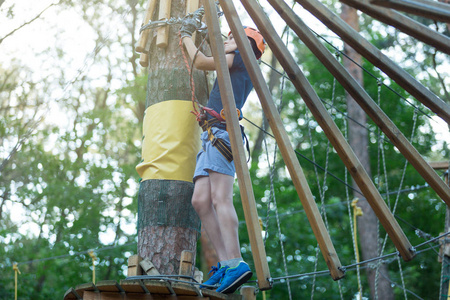 快乐可爱的小男孩穿着蓝色T恤和橙色头盔，在冒险公园玩，拿着绳子和爬木楼梯。 积极的生活方式概念
