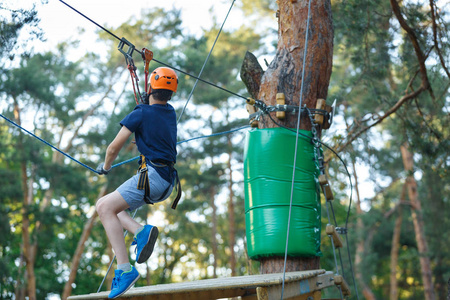 快乐可爱的小男孩穿着蓝色T恤和头盔，在冒险公园玩，拿着绳子和爬木楼梯。 积极的生活方式概念