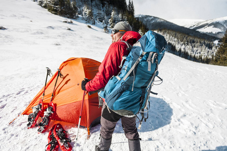 冬天在帐篷附近的人。 极端条件下的游客。 雪鞋徒步旅行杆和背包。 在冬天的山上露营。 雪地里的橙色帐篷。