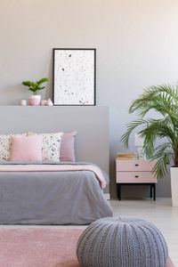 灰色的头巾旁边的床与垫子在现代卧室内部与海报和植物。 真实照片
