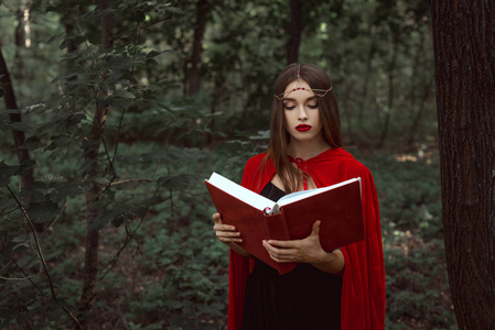 迷人的神秘女人穿着红色斗篷和花环在森林里读魔法书