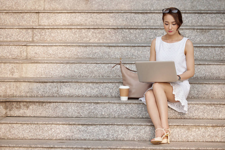 优雅的亚洲女人，穿着白色连衣裙，坐在楼梯上，拿着笔记本电脑和一杯咖啡