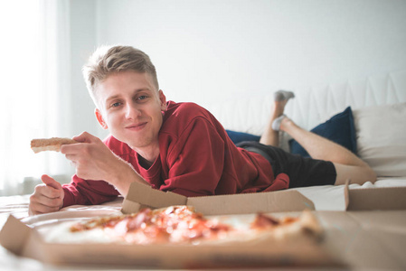 穿着红色运动衫的快乐迷人的年轻人躺在披萨盒床上舒适的房间里。 积极的青少年手里拿着一块比萨饼，看着相机微笑。