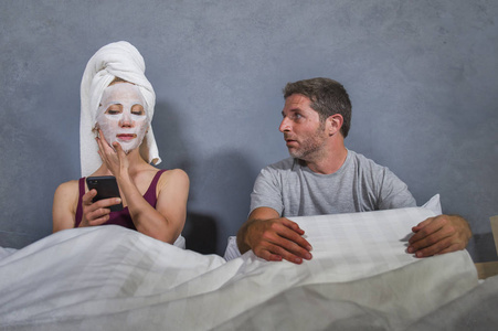 有趣的生活方式肖像古怪古怪的家庭主妇化妆面膜和毛巾使用手机在床上和丈夫绝望的脸表情在男人女人关系的概念