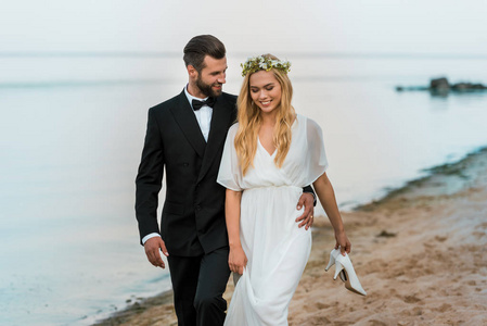 深情的新婚夫妇拥抱和漫步在海滩新娘手里拿着高跟鞋