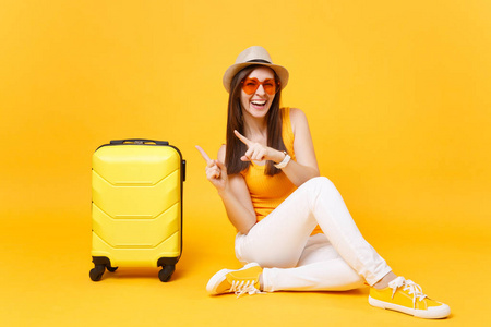 旅行者旅游妇女戴着夏季休闲服装帽，坐在黄色橙色背景上的手提箱上。 女乘客在周末出国旅行，度假。 航空旅行概念