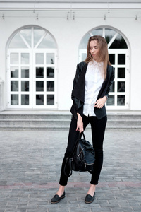 时尚漂亮的年轻女人穿着时尚的黑色皮夹克，白色衬衫，黑色裤子，背着黑色背包。
