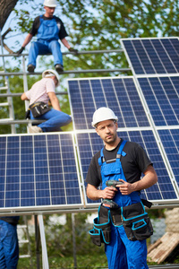 成功的工程师技师的肖像与电气螺丝刀站在未完成的高外部太阳能电池板照片Voltaic系统与团队的工人在高平台背景。