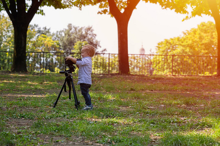 两岁的男孩是摄影师。 孩子拿着相机在三脚架上拍照。 小孩子穿着衬衫和牛仔裤在公园里玩。