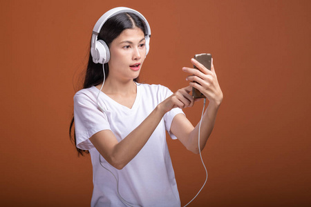 女人听音乐用电话隔离在橙色背景上。 年轻的亚洲女人带着手机听音乐浏览音乐。 时尚生活方式概念