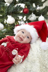 可爱的小宝宝穿着红色圣诞老人服装躺在装饰圣诞树附近
