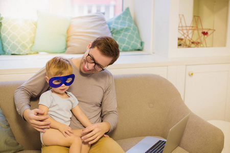 年轻快乐的父亲和他的小儿子坐在房间里的沙发上戴着面具