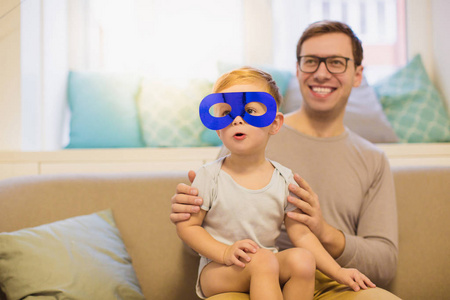 年轻的微笑的父亲和他的小儿子坐在房间里的沙发上戴着面具