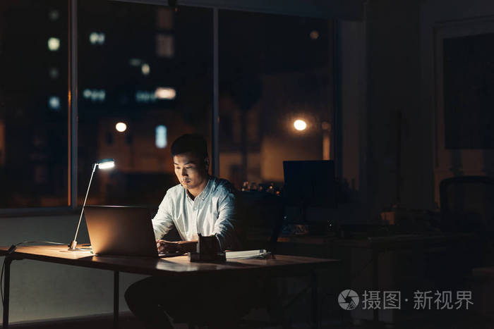 年轻的亚洲商人在笔记本电脑上工作,晚上坐在办公桌前一间黑暗的办公