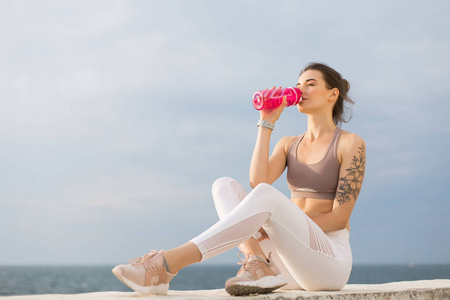 年轻漂亮的女士，穿着运动上衣和白色紧身裤，手持粉红色运动瓶，饮用水，背景海景。