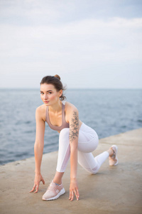 美丽的女孩在运动的顶部和白色的腿练习瑜伽与海景的背景。 一位年轻的女士梦幻般地看着在海边伸展的镜头