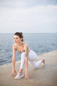 美丽的女孩在运动的顶部和白色的腿练习瑜伽与海景的背景。 年轻女子若有所思地看着海边的训练
