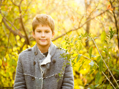 可爱的，年轻的，时尚的小男孩的肖像，穿着灰色的外套在树上。在明亮的黄色秋天的大自然中行走。家庭户外活动。