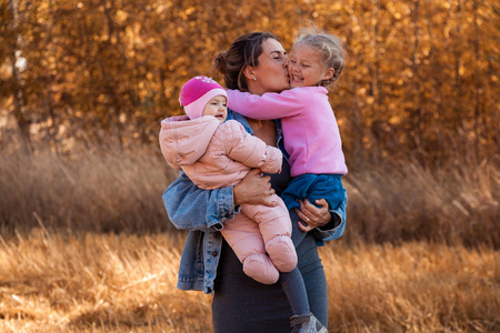 一位年轻的母亲抱着她的女儿，在秋天田野景观和黄色树木的背景下，摆姿势和与她们交谈，享受大自然的乐趣。