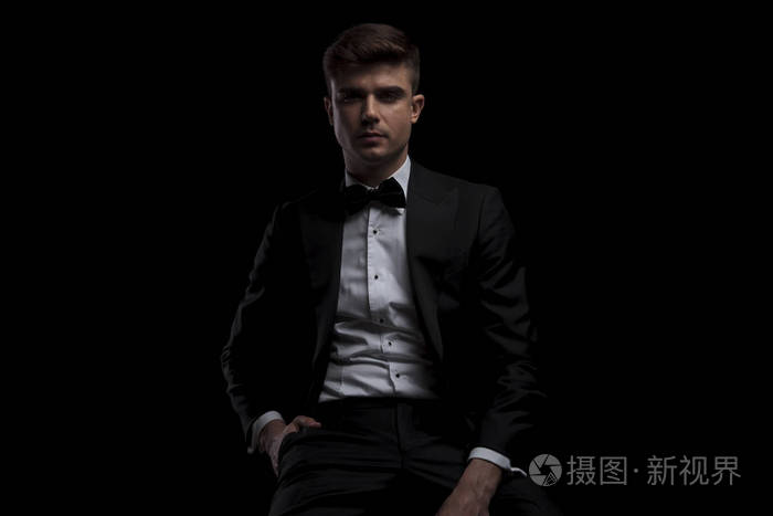 一个穿着黑色西装,手插口袋,坐在黑色背景上的轻松男人的肖像照片-正