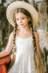 一个可爱的小女孩，长着一头金色的长发，穿着白色的沙拉帆，戴着一顶草帽，坐着一把藤椅和一根芦苇装饰
