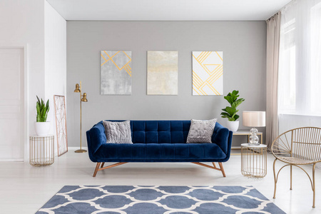 一个优雅的深蓝色沙发在一个明亮的客厅内部，金色的金属边桌和三幅画在灰色的墙上。 真正的照片。