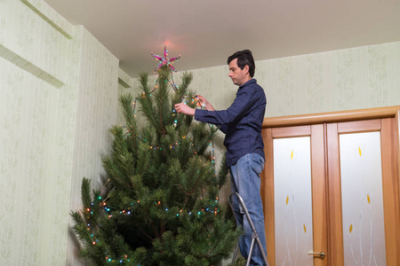 英俊的中年男子装饰了一棵圣诞树，一颗星星和一个五颜六色的灯泡花环站在客厅内部的梯子上。 新年假期概念舒适的家