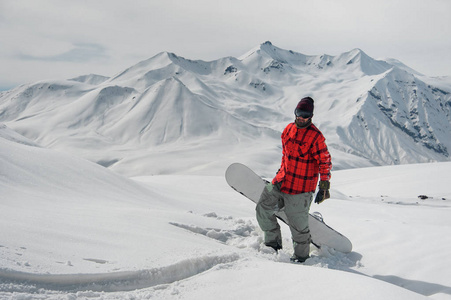 年轻的男性滑雪者站在山坡上，双手拿着滑雪板，站在山的背景和晴朗的天空上