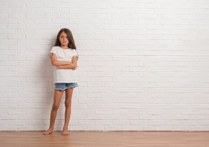 年轻的西班牙裔儿童站在白色砖墙怀疑和紧张的不赞成的表情在脸上交叉的手臂。 消极的人。