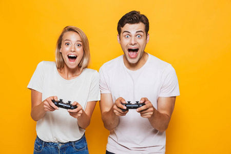 照片快乐兴奋的年轻情侣站在黄色的墙壁背景上玩游戏与操纵杆。
