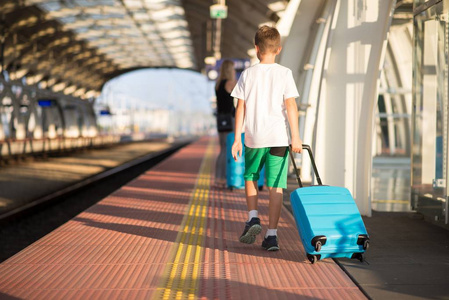 母亲和儿子带着蓝色行李箱在火车站站台上行走，等待火车