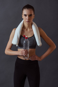 穿着运动服的漂亮女孩拿着一瓶水，看着灰色背景的相机