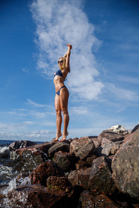 美丽的金发女人穿着泳装站在岩石海滩上，白天