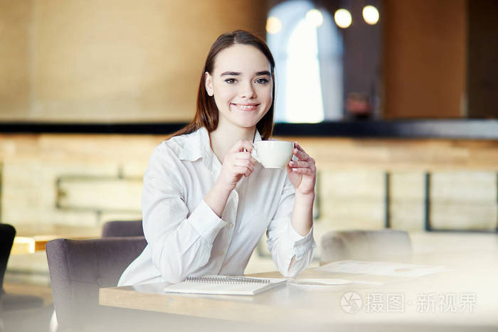 美丽的年轻女商人坐在咖啡桌上,手里拿着咖啡杯,看着相机,微笑着