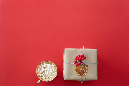 圣诞礼物是用手工纸包装的，上面装饰着一根红色的振动树枝，红色背景上有一杯可可和棉花糖。 顶部视图复制空间