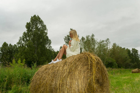 一个穿着白色针织羊毛夹克的漂亮金发女人坐在一大堆干干草上，这些干草是在夏天或秋天收集的，在半个转弯的绿色草地上收集的