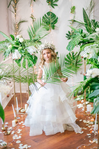 可爱的小女孩，金色的卷发，穿着白色的婚纱，戴着热带树叶白色玫瑰和兰花装饰的花圈