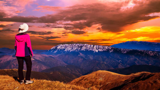 山岭上空令人叹为观止的日落。 女人坐在山顶上，看着她面前群山上的夕阳。