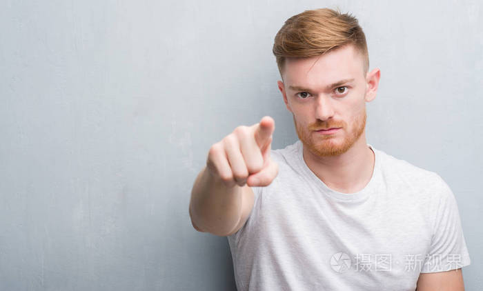 年轻的红发男子在灰色的灰色墙壁上用手指指向相机和你的手标志积极和