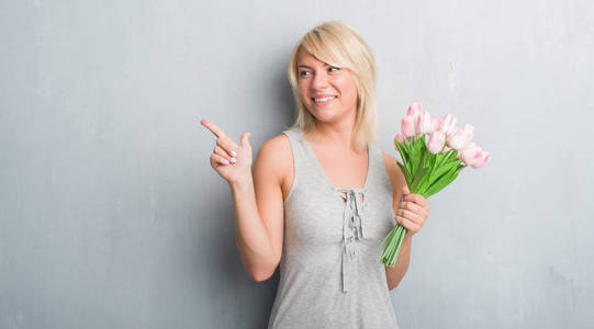白种人成年妇女在灰色的格朗格墙上抱着粉红色的花，非常高兴地用手和手指指向一边