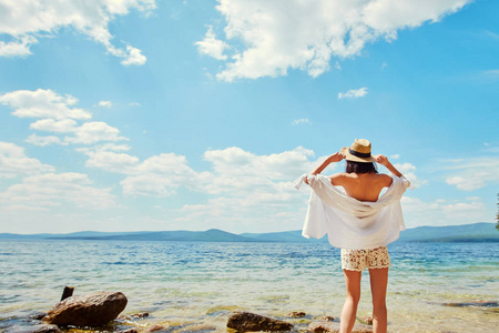 一个穿着白色衬衫和草帽的苗条女孩背着她站在海湾的岸边。