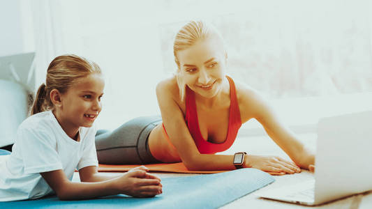 妈妈和女儿盯着笔记本电脑屏幕。 休息一下。 一起放松。 在家健身。 假日休闲。 身体平衡。 躺在健身房的地毯上。 家庭关系。 晴
