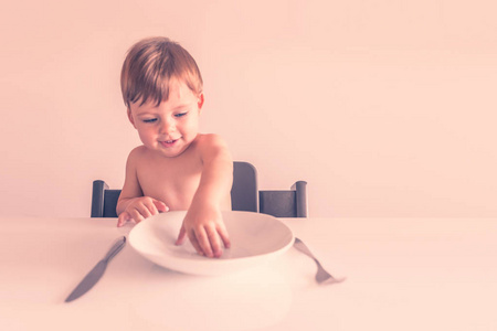 可爱的金发小男孩坐在桌子旁，手里拿着一把空的白色盘子叉子和刀子