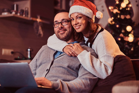 一对可爱的夫妇的肖像与笔记本电脑迷人的女人在圣诞老人的帽子拥抱她的男人和看相机