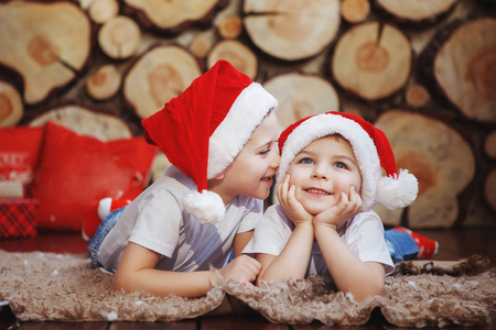 两个戴着圣诞帽的兄弟男孩躺在新年树附近的地毯上，在砍树的背景下笑着。