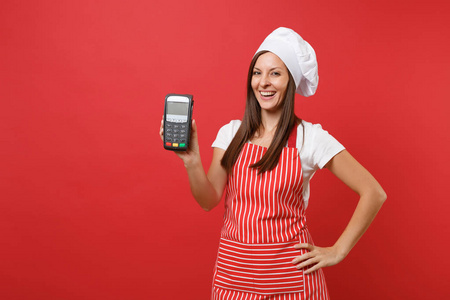 家庭主妇，女厨师或面包师，穿条纹围裙，T恤，厨师帽子，隔离在红墙背景。 女性手持无线银行支付终端NFC设备。 模拟复制空间概念