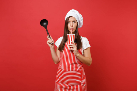 家庭主妇女厨师或面包师穿条纹围裙，白色T恤，烤厨师帽子隔离在红墙背景上。 女人拿着汤，黑瓢，塑料杯可乐。 模拟复制空间概念