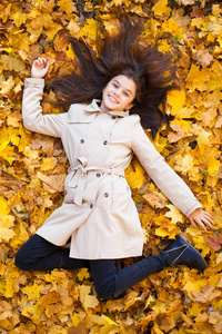秋天的公园里，一个年轻漂亮的小女孩穿着米色外套，躺在上面的黄叶上